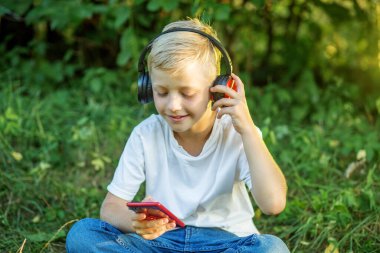 Child listens to music. Blond boy uses headphones and smart phone. Preteen boy listening to song or audio book.