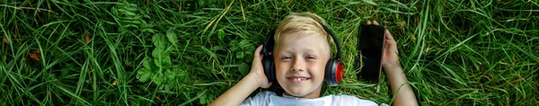 Happy child listens to music and smiles. Blond boy uses headphones and smart phone, lies in grass and rests. Website banner