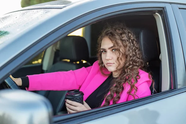 Young woman driving car. Portrait of curly woman in car looking out window and drinking hot coffee. Pink jacket. Business concept.