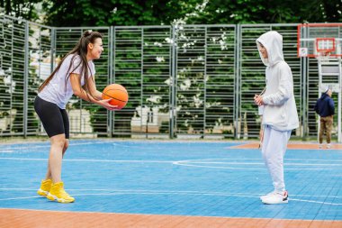 Kız ve küçük kardeşi, genç kız, açık gökyüzünün altında, modern basketbol sahasında basketbol oynuyorlar. Spor anlayışı, hobiler ve sağlıklı yaşam tarzı.