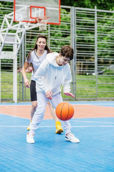 Kız ve küçük kardeşi, genç kız, açık gökyüzünün altında, modern basketbol sahasında basketbol oynuyorlar. Spor anlayışı, hobiler ve sağlıklı yaşam tarzı.