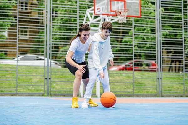 Girl and her younger brother, teenager, play basketball on modern basketball court under open sky. Concept of sports, hobbies and healthy lifestyle.