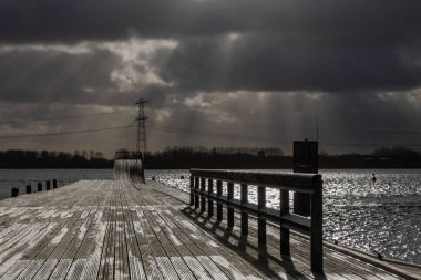 Landscape with stormy clouds and sunrays over a wooden pier with black birds facing the wind and a dark lake. In the distance, there are high poplar trees together with an electricity mast and cables