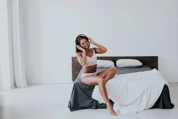 Bela mulher em lingerie ouvindo música em fones de ouvido na cama no quarto — Fotografia de Stock