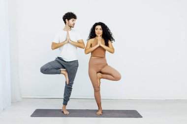 Bir kadın ve bir erkek, beyaz bir odada çift yoga yapıyorlar.