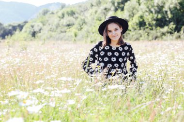 Elbise ve şapka giymiş güzel bir kadın papatya tarlasında yürüyor.