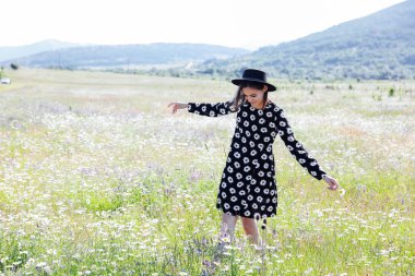 Elbise ve şapka giymiş güzel bir kadın papatya tarlasında yürüyor.