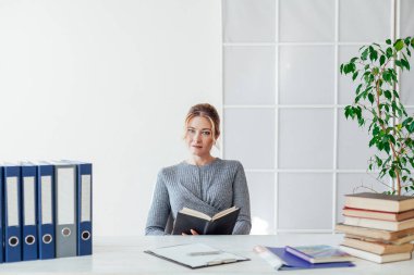 Takım elbiseli bir iş kadını, elinde kitaplarla bir ofis masasında oturuyor.