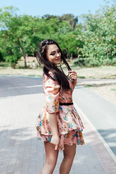 Piękna brunetka kobieta w sukience z kwiatami spacery wzdłuż ulicy i parku — Zdjęcie stockowe