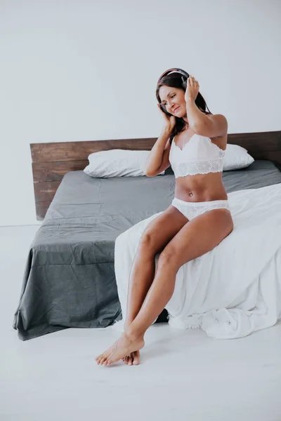 Bela mulher em lingerie ouvindo música com fones de ouvido no quarto na cama — Fotografia de Stock