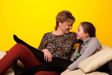 Anne ve kız, sarı arka planda, siyah bir kanepede.