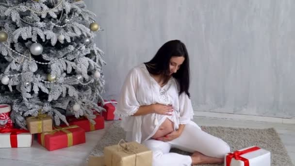 Schöne schwangere Frau sitzt am Weihnachtsbaum mit Neujahrsgeschenken — Stockvideo