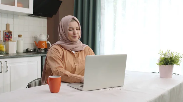 Woman in hijab working remotely on laptop in kitchen. Young woman using laptop in kitchen drinking coffee while working.