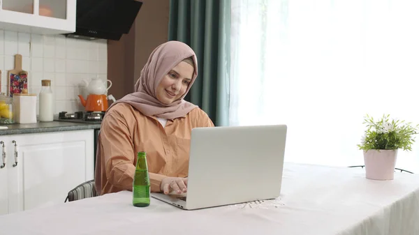 Hijab dressed woman working on laptop in kitchen, freelancer remote business messaging message take a break, drink natural mineral water, take a sip of health care, prevent dehydration. Daily routine