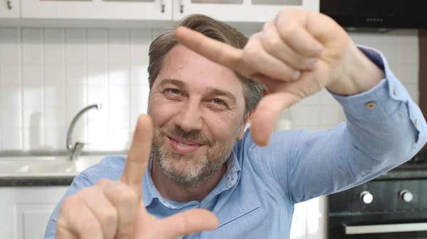 Man sitting in kitchen making frame gesture. He is looking at the camera and making a camera frame with his hands.