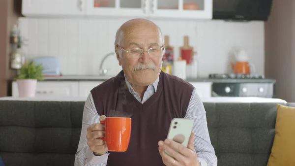 Old man watching funny video on his smartphone while drinking coffee at home. The man is laughing at the sms or video he sees on his hairy phone. The concept of technology use in the elderly.