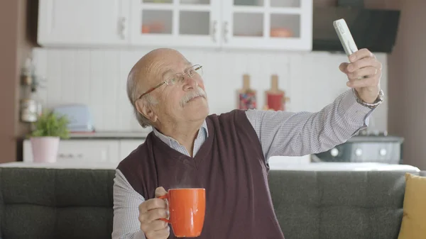Happy 70s male user making video call online while drinking coffee, looking at screen sitting on sofa at home, taking selfie, old grandfather learning to use modern technology.