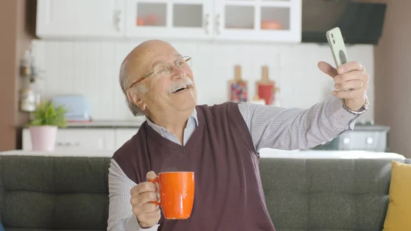 Happy 70s male user making video call online while drinking coffee, looking at screen sitting on sofa at home, taking selfie, old grandfather learning to use modern technology.