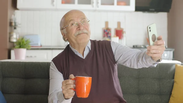 Happy 70s male user making video call online while drinking coffee, looking at screen sitting on sofa at home, taking selfie, old grandfather learning to use modern technology.