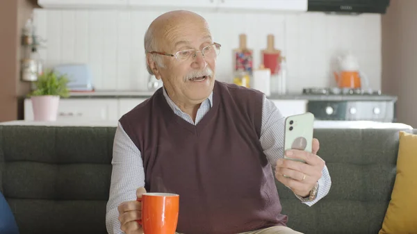那位老人在智能手机上拨号 在家的时候给手机发短信 80多岁的老人在智能手机上看手机应用程序 短信和新闻 他对着镜头笑 — 图库照片