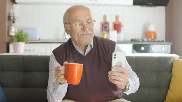 老人一边在家里喝咖啡 一边在智能手机上看滑稽的视频 这个人正在嘲笑他在毛茸茸的手机上看到的短信或视频 老年人使用技术的概念 — 图库照片