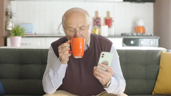老人一边在家里喝咖啡 一边在智能手机上看滑稽的视频 这个人正在嘲笑他在毛茸茸的手机上看到的短信或视频 老年人使用技术的概念 — 图库照片