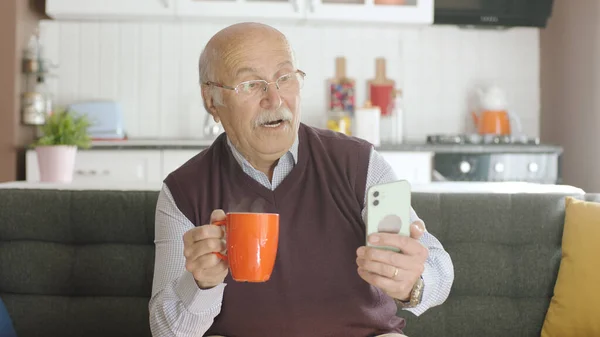 Old man watching funny video on his smartphone while drinking coffee at home. The man is laughing at the sms or video he sees on his hairy phone. The concept of technology use in the elderly.