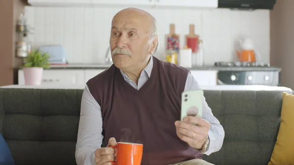 Old man watching funny video on his smartphone while drinking coffee at home. The man laughs at the sms or video he sees on his smartphone and looks at the empty advertising space.