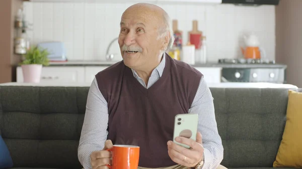 Old man watching funny video on his smartphone while drinking coffee at home. The man laughs at the sms or video he sees on his smartphone and looks at the empty advertising space.