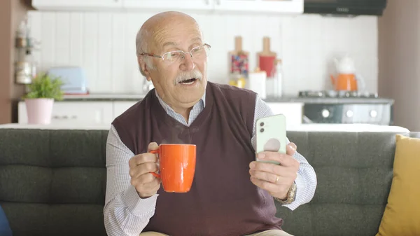 Old man watching funny video on his smartphone while drinking coffee at home. The man laughs at the sms or video he sees on his smartphone and is very surprised at what he sees.