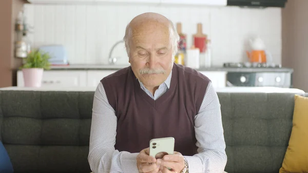 Old man watching funny video on his smartphone while drinking coffee at home. The man is laughing at the sms or video he sees on his hairy phone. The concept of technology use in the elderly.