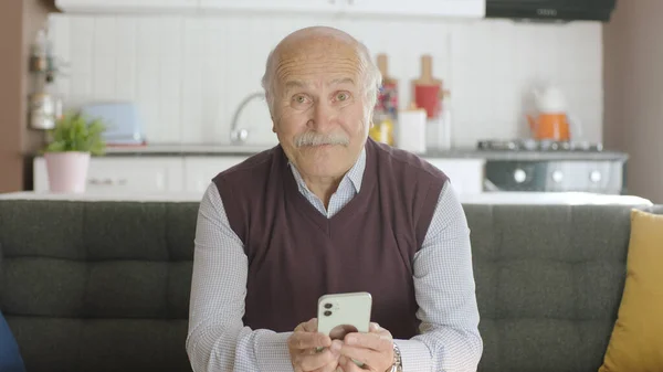 The old man dials the number on the smartphone. Sends sms to mobile phone while at home. 80s old man looking at mobile apps, sms and news on his smartphone. He is smiling at the camera.