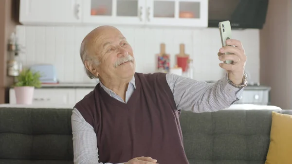 Happy 70s male user making video call online while drinking coffee, looking at screen sitting on sofa at home, taking selfie, old grandfather learning to use modern technology.