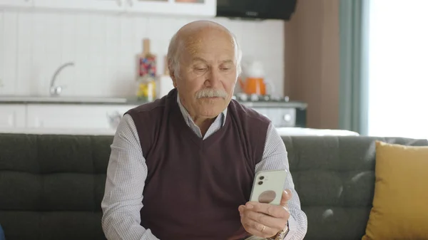 The old man dials the number on the smartphone. Old man sends sms to mobile phone while home. 80s old man looking at mobile apps, sms and news on his smartphone.