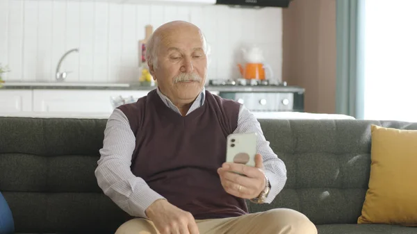 The old man dials the number on the smartphone. Old man sends sms to mobile phone while home. 80s old man looking at mobile apps, sms and news on his smartphone.