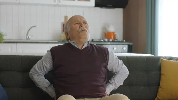 Elderly man sitting on sofa in living room at home suffering from chronic low back and back pain. Back and lower back pain in old age, health problems concept.