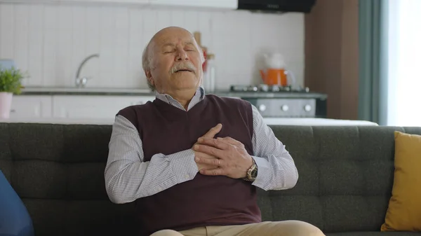 Feeling of discomfort, risk of heart attack. Elderly man feeling chest pain while sitting on sofa at home having difficulty breathing. He touches his chest with his hand.