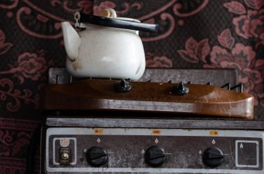 Aceleyle atılmış, eski bir gaz ocağı, örümcek ağlarıyla kaplı eski bir su ısıtıcısı, küçük bir su ısıtıcısı, desenli bir halı, terk edilmiş bir mutfak.