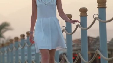 Akşamları sahilde uzun bir rıhtımda yürüyüşe çıkan beyaz elbiseli genç bir kadın. Tatil ve seyahat kavramı.