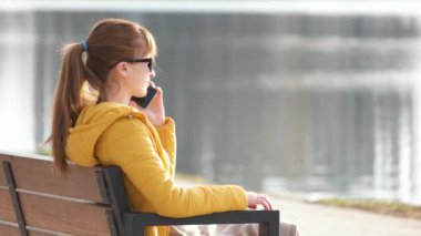Parktaki bankta oturan genç ve güzel bir kadın sıcak bir sonbahar akşamında dışarıda akıllı telefonuyla konuşuyor..