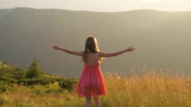 Kırmızı elbiseli genç mutlu kadın yolcunun arka görüntüsü. Çimenli bir yamaçta duruyor. Yazın rüzgarlı bir akşamda, kollarını açıp güneşin batışının keyfini çıkarıyorlar..