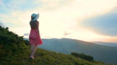 Kırmızı elbiseli genç mutlu kadın gezgin yaz dağlarında rüzgarlı bir akşamda çimenli yamaçta duruyor gün batımında doğanın tadını çıkarıyor..