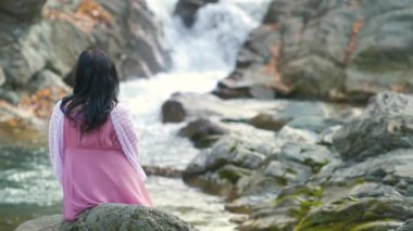 Uzun, şık bir gece elbisesi giymiş, siyah saçlı bir kadın hızlı akan suyla birlikte küçük bir dağ nehrinin kıyısındaki bir taşın üzerinde oturuyor..