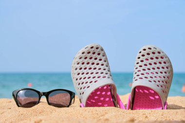 Sıcak, güneşli bir günde tropik deniz kıyısındaki kumlu kumsalda takunya ayakkabıları ve siyah koruyucu güneş gözlükleri. Yaz tatili kavramı.