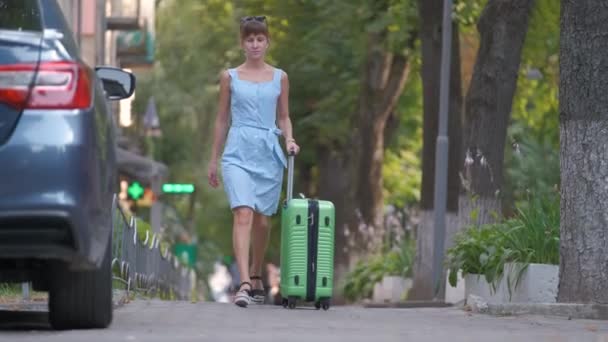 Молодая женщина, идущая по улице, кладет зеленый чемодан в машину. Концепция путешествий и отдыха — стоковое видео