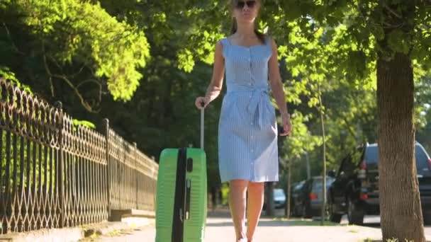 Jeune femme marchant sur le trottoir de la rue de la ville avec valise verte le jour de l'été. Concept de voyage et de vacances. — Video