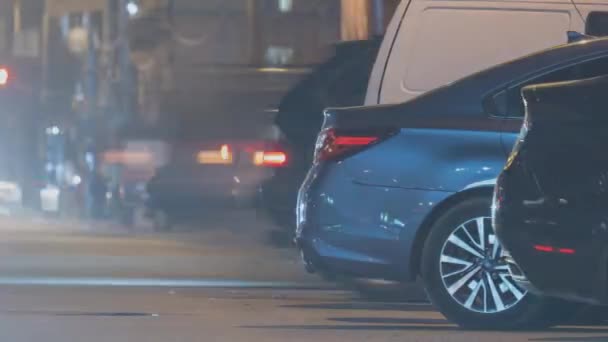 Zblízka zaparkovaných aut na silnici v noci s rozmazaným výhledem na semafory jedoucích vozidel na ulici — Stock video