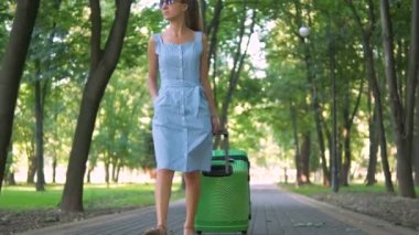 Genç bir kadının yaz günü yeşil bavuluyla şehir kaldırımında yürümesi. Seyahat ve tatil kavramı