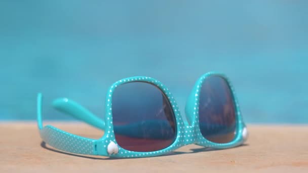 Primer plano de gafas de sol azules en el lado de la piscina en un día cálido y soleado. Concepto vacaciones de verano. — Vídeos de Stock