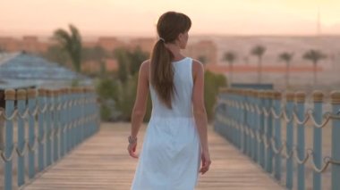 Beyaz elbiseli genç bir kadın uzun rıhtımda yürüyüşe çıkıyor. Akşamları dağ zirveleri siluetiyle. Tatil ve seyahat kavramı.